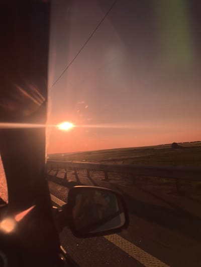 The image depicts a sunset viewed from inside a vehicle, likely on a highway or rural road. The sun, a bright orb with a lens flare, is positioned low on the horizon, casting a warm, orange glow across the sky and the landscape. The sky is a gradient of pink and orange, with a faint hint of green in the upper right corner, possibly from atmospheric scattering or a lens effect.

To the right of the sun, a vast, rolling landscape of green fields stretches to the horizon, dotted with a few scattered trees. A metal guardrail runs along the side of the road, separating the vehicle from the open fields. The road surface appears to be asphalt, with a yellow and white striped marking visible in the foreground.

In the foreground, the dark silhouette of the vehicle's A-pillar and side mirror dominates the left side of the frame. The side mirror reflects a partial view of the interior of the vehicle, showing a person's head and shoulders, possibly the driver or a passenger. The overall impression is one of a serene and picturesque journey during golden hour.

There is no indication of the city of Yumorga, Russia, or any specific geographical features that would place the image there.