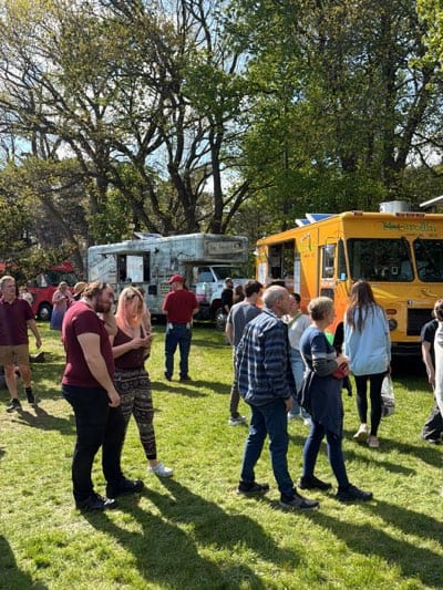The image depicts an outdoor event in Rochester, United States, likely a food truck rally. The location appears to be a grassy area or park, with trees providing a backdrop. Two food trucks are visible: one yellow, labeled "Macarollin", and another with a distressed or aged design. A variety of people are present. Some stand near the food trucks, possibly waiting in line or socializing, while others appear to be walking through the area. They are dressed in casual clothing, suggesting a relaxed atmosphere. The scene is well-lit, indicating it is daytime.