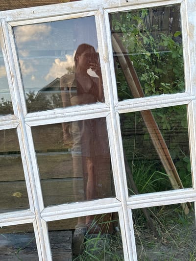 In the city of Кривандино, Russia, an old, whitewashed wooden window frame with multiple panes is propped up against a weathered wooden fence. The glass panes reflect the surrounding environment and a person holding a smartphone, taking a selfie. The reflection shows a person's upper body and head, with a dark top and blonde hair. They are holding a phone in their right hand, angled to capture the reflection. Behind the person, the reflection reveals a blue sky with white clouds and the roofline of a building. Another pane of glass shows a dense green garden with leafy plants and a wooden structure. The overall scene depicts an outdoor setting, likely a backyard or garden area, with elements of neglect and rustic charm. The angle of the window frame suggests it might be leaning or resting against something. The lighting indicates it is daytime, with sunlight illuminating the scene.