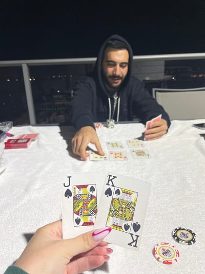The image depicts a nighttime card game, likely poker, occurring outdoors on what appears to be a balcony overlooking a city skyline. The specific location within Yavne, Israel, is not discernible from the visual information provided.
Two individuals are present. In the foreground, a female hand with almond-shaped nails featuring pink and white French tips holds two playing cards: the Jack of Spades and the King of Spades. Directly opposite, across a white-covered table, an adult male with dark hair and a beard, wearing a dark hooded sweatshirt, is seated. His right hand is positioned over one of the community cards on the table, while his left hand holds several red-backed playing cards.
On the white, textured tablecloth, four community cards are visible: a partially obscured Queen of Clubs, a King of Hearts, an Ace of Hearts, and a 5 of Diamonds. To the left, a red-backed deck of cards, a clear glass ashtray, and a pink marker or pen are present. To the right, near the foreground hand, two "6 POKER" branded poker chips (one black and white, one red and white) are on the table. A dark-colored smartphone is also visible on the right side of the table, near the male participant. The background shows a dark night sky above a cityscape illuminated by numerous lights. A light-colored railing or barrier is visible between the table and the distant city lights. The scene is illuminated by an unseen artificial light source, casting shadows on the tablecloth.
