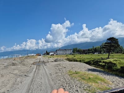 A low-angle, wide shot captures a sandy, undeveloped beach area leading towards the ocean in Adzhiryuara, Abkhazia. In the foreground, a sandy track with tire marks suggests recent vehicle activity, with a small puddle visible on the left. Piles of sand and some construction debris, including wooden planks, are scattered across the beach. In the mid-ground, a resort-style building with multiple balconies and archways stands prominently, surrounded by green trees. To the right of the building, a large coniferous tree stands tall. Further in the distance, rolling mountains form the backdrop under a bright blue sky dotted with fluffy white clouds. Several white umbrellas are set up on the beach near the water's edge, where a few people are visible. A white caravan or recreational vehicle is parked near the buildings. The overall impression is one of a coastal area undergoing some form of development or renovation, with a contrast between the natural landscape and the constructed elements.