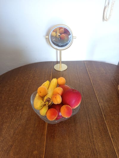 The image shows a still life on a wooden table. In the foreground, a clear glass bowl holds a selection of fruits: several ripe bananas, multiple apricots, and one bright red apple. Behind the fruit bowl, a gold-colored vanity mirror with LED lighting stands on a thin gold stand. The mirror reflects some of the fruit. The background is a plain white wall. A coiled white cord hangs from the top right of the wall. There is no indication of the city of Nancy, France, or any other location. The scene is static and shows no people or actions.