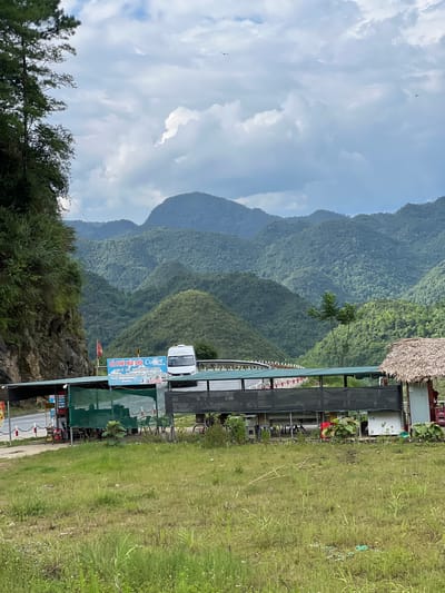 The image depicts a roadside rest stop in Quản Bạ Commune, Vietnam, nestled amidst a dramatic landscape of lush green mountains under a cloudy sky. A white van is parked in front of a covered seating area, which appears to be a cafe or small eatery. A sign in Vietnamese, featuring the words "CA PHE NUI DOI" (Mountain Coffee), is prominently displayed. Tables and chairs are set up under the canopy, and some are covered with green netting. To the right, a small structure with a thatched roof stands, possibly a souvenir shop or additional seating. The foreground is dominated by a grassy field, with a dirt path and a road visible to the left. The overall impression is one of a scenic, rural stopping point along a mountainous route.