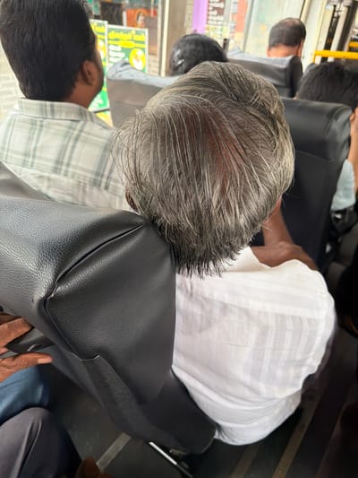 A man with graying hair is seated on a bus, his head leaning against a black bag. He is wearing a white collared shirt. To his left, another man in a checkered shirt is visible. The bus interior, with black seats, is apparent. The scene takes place on a bus in Madirimangalam, India.
