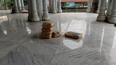 The image shows an outdoor area with a polished marble floor and large white pillars. In the foreground, there are several bags of cement. A stack of five bags is on the left, with one additional bag lying flat to the right of the stack. A smaller, torn bag is also present next to the stack of cement bags. In the background, there are more pillars and a railing. Beyond the railing, a building with a red roof is visible, along with some trees and a sign that appears to be in Indonesian. The scene suggests a construction or renovation site within a sheltered area, possibly a temple or a pavilion, in Beunyot, Indonesia.