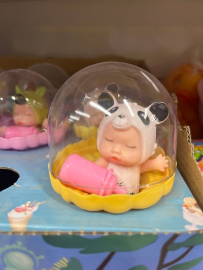 In the city of Ivanivka, Ukraine, a close-up shot focuses on two infant-like toys displayed in clear plastic domes. The toy in the foreground is dressed in a white outfit with a panda-themed hood, complete with black ears and facial markings. It lies on its back in a yellow, petal-shaped base, holding a pink bottle. Its eyes are closed, and its mouth is slightly open, giving the impression of sleep. Behind it, another similar toy is visible, wearing a yellow hood and resting in a pink base, also holding a pink bottle. The toys are placed on a blue surface with illustrations of ice cream cones. The background is blurred, showing shelves and other merchandise, suggesting a retail environment.