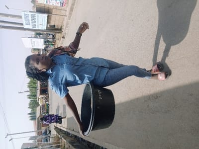 The image depicts an outdoor street scene within Mumbwa, Zambia. A female individual, exhibiting dark skin and braided hair, stands on a light grey paved surface under bright daylight. She is attired in a light blue short-sleeved denim shirt, blue jeans, and pink slip-on sandals. Her left hand rests on the upper rim of a large, black, cylindrical plastic container situated to her left. Her right arm is bent and slightly raised, with a dark wristband visible on her wrist. Prominent shadows of the individual and the container are cast on the ground to her right. In the middle ground, further along the paved road, a second individual in a dark purple patterned garment walks away from the camera's perspective. The background features several utility poles, two billboards (the upper one clearly displaying "BELIEVERS' HIGH SCHOOL"), various single-story buildings suggestive of commercial or residential use, and distant trees. A shallow ditch or gutter runs alongside the paved surface on the left.