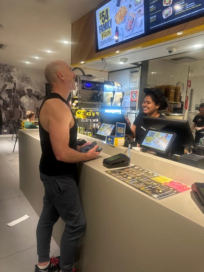 The image shows an indoor scene at a fast-food restaurant, likely Taco Del Mar, given the visible text. A male customer, bald with a muscular build, is standing at a counter, looking upwards. He is wearing a black tank top and dark grey sweatpants. To his right, a female employee in a black t-shirt and black cap is smiling and interacting with a customer at the register. Several digital menu boards and a payment terminal are visible on the counter. Behind the employee, a large menu board displays various food items and prices, including "FAMILY FIESTA $54" and "FRIENDS FIESTA $30". Another menu board also shows "FRIENDS FIESTA $30". Various drink dispensers and kitchen equipment are visible in the background. The restaurant appears to be clean and well-lit, with modern decor. A black and white mural of people playing musical instruments adorns the wall behind the customer. The overall atmosphere seems casual and friendly. The presence of digital menus and payment terminals suggests it is daytime. The visible text, including the restaurant name and menu items, provides specific details about the establishment and its offerings.