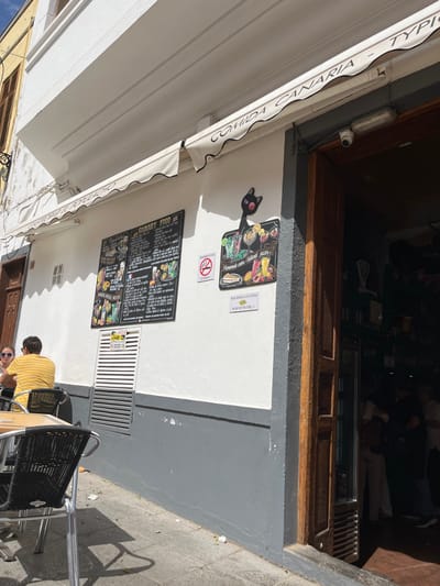 The image shows the exterior of a restaurant in Agüimes, Spain, located at coordinates 27.907, -15.447. The restaurant has a white facade with a gray stripe running along the base. A black cat silhouette is affixed to the wall above a chalkboard menu advertising "Canary Food" with a variety of options. A brown awning overhangs the restaurant entrance, with the words "Comida Canaria - Tipis" written in black and white lettering. The awning extends over a wooden doorway that leads inside, where there are several people standing near a refrigerator, likely staff or customers. A "No Smoking" sign is visible next to the menu, and a sign that reads "No se Molesten" ("Don't Disturb") is attached below it.  A man in a yellow shirt is sitting at a table outside the restaurant, looking towards the doorway.  A woman in a white shirt is sitting to his left, her back to the camera.  A black chair is in front of her, and there is a black table beside her, with a chair partially visible behind her. There are more chairs along the restaurant's exterior wall, and the ground is covered in gravel and small stones. The sky is visible in the upper right of the image.