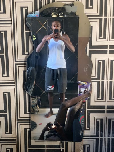 An individual with dark skin, wearing a white tank top and dark grey shorts with a reversed "RHUDE" logo, stands barefoot, capturing a mirror selfie indoors in Osara Gada, Nigeria. The subject holds a black smartphone with both hands at chest level, facing the device, with eyes partially or fully closed. The reflection is captured in an irregularly shaped mirror. The upper torso and head are clearly visible, while the lower portion of the mirror reflects the individual's bare legs, appearing crossed at the ankles, and hands holding another phone, as if seated or crouching.
The background visible in the mirror's reflection consists of a dark-colored wardrobe or cabinet. On its top surface, a black backpack, a pair of light-colored sneakers, and a small white and red box are present. To the left of the reflected person, a black oscillating fan is visible. To the right, a purple multi-tiered plastic caddy containing various small items is situated. The floor visible in the reflection is tiled white, with a small patterned bag and a red cylindrical object on its surface. The wall behind the mirror is adorned with a distinctive black and white geometric pattern.