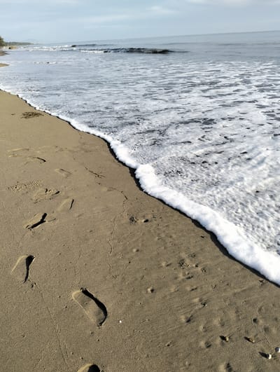 The image depicts a beach scene in Sagoe, Indonesia. The foreground shows a sandy beach with visible footprints. White foamy waves are washing onto the shore. The ocean extends into the background, where faint waves can be seen. The sky is partly cloudy.