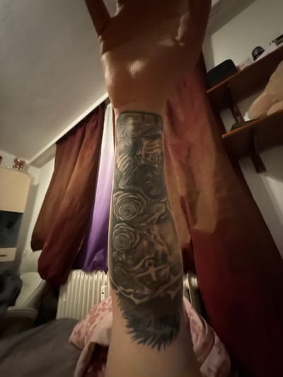 A close-up, angled shot focuses on a person's forearm bearing a grayscale tattoo design. The tattoo appears to depict a combination of roses, possibly a lion, and other indiscernible elements in a detailed, shaded style. The arm is positioned horizontally across the frame, with blurred background elements suggesting an indoor setting. These elements include a variety of fabrics in shades of red, brown, purple, and pink, along with glimpses of furniture and household items, such as a chair and a radiator. The specific location in Lüneburg, Germany, cannot be determined from the visual information provided.