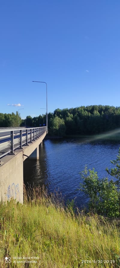 The image depicts a concrete bridge stretching over a body of water, likely a river or a lake, in Hartola, Finland. The bridge has a wide concrete walkway bordered by metal railings on one side, and on the other, it slopes down towards a lower level with more metal structures. Streetlights are visible along the bridge. The water is dark blue and appears to be slightly rippled, reflecting the clear blue sky above.
On the left side of the image, a dense line of lush green trees forms a backdrop, creating a natural boundary against the sky. To the right, tall, dry grass and foliage dominate the foreground, adding a touch of wildness to the scene. A small section of a tree with green leaves is visible at the top right, slightly obscuring the view of the water.
The time of day appears to be late afternoon or early evening, suggested by the lighting and the presence of the date stamp "2026/6/30 20:29". The weather seems clear and sunny, indicated by the bright blue sky with a few wispy clouds. The overall atmosphere is serene and natural, highlighting the integration of the man-made structure with the surrounding Finnish landscape. There is some graffiti on the concrete of the bridge. The text "REDMI NOTE 9 PRO AI QUAD CAMERA" is present in the bottom right corner of the image, indicating the device used to capture the photograph.