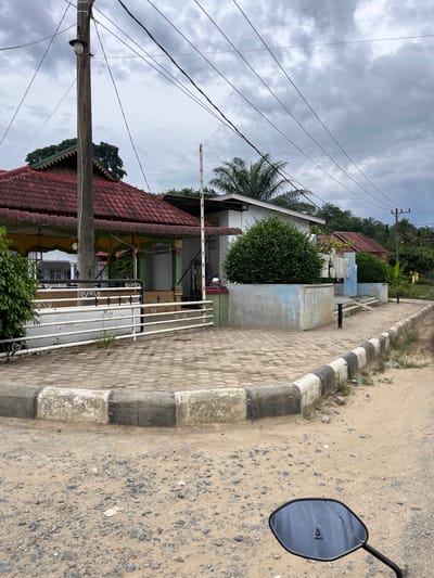 The image depicts a street scene in Rantau Pauh, Indonesia. In the foreground, the reflection of a motorcycle's rearview mirror is visible on a sandy and gravelly road. The road has a distinct curb with alternating black and white painted sections. Adjacent to the road is a paved area with hexagonal tiles. Beyond the paved area, a white metal fence encloses a property with a building. The building has a red tiled roof with yellow and green trim around its eaves and supporting columns. A tall utility pole with electrical wires runs overhead. Lush green vegetation, including a large bush and trees, surrounds the property and extends into the background, where more trees and palm trees are visible under a cloudy sky.