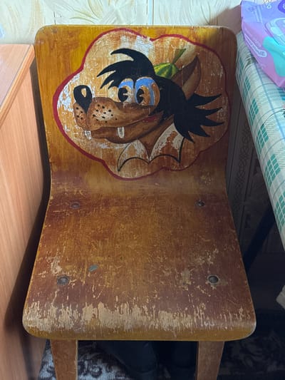 A close-up shot focuses on an old, wooden stool with a cartoonish depiction of a wolf painted on its seat. The stool appears worn, with visible scratches and chipped paint. The surrounding environment includes a tiled wall, a wooden surface, and what appears to be household items above and behind the stool. The location is specified as Chuprino, Russia.