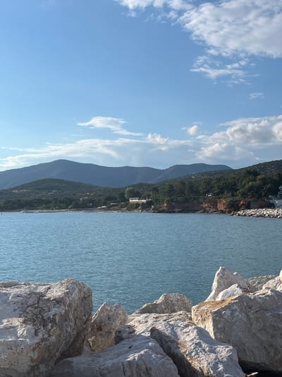 A vertical, eye-level outdoor shot shows a picturesque coastal scene under a bright blue sky dotted with white clouds. In the foreground, large, light-colored rocks, some with rough textures, are piled up, suggesting a breakwater or rocky shoreline. Beyond these rocks, a calm, blue body of water stretches out, its surface rippled by a gentle breeze. The middle ground reveals a lush, green coastline with trees and some buildings visible. In the background, a series of rolling hills and mountains, covered in vegetation, form a dramatic backdrop against the sky. The overall impression is one of natural beauty and tranquility. The location is the coast of Potós, Greece.