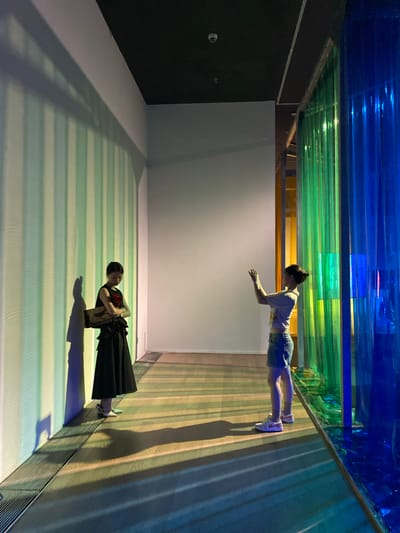 Two individuals are positioned within a brightly lit interior space, possibly an art installation or gallery, with a dark ceiling. On the left, a person in a black dress with a red floral accent and white shoes stands with their arms crossed, holding a beige clutch. They are illuminated by light casting vertical stripes of green and white on the wall behind them. To the right, another person wearing a white t-shirt, denim skirt, and white sneakers stands with their hands raised, appearing to interact with something. This individual is positioned near a translucent wall composed of vertical strips of blue and green. The floor is a wooden surface with prominent shadows cast by the lighting and the people present. The overall environment suggests an artistic or exhibition setting, possibly within Jinniu District, China, given the provided context.