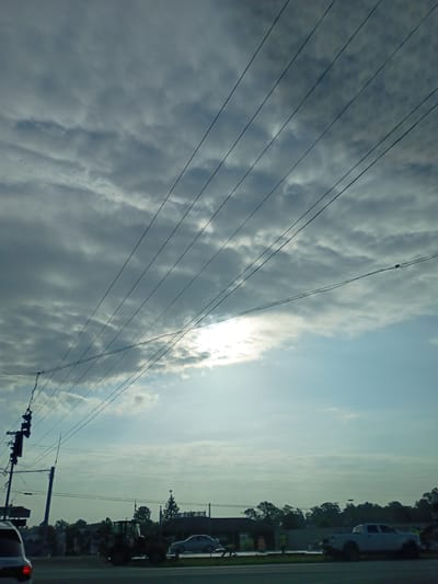 A medium-shot, eye-level, outdoor, daylight, wide-angle photograph shows a cloudy sky with several power lines running diagonally across the frame from left to right. The sun is partially visible through a break in the clouds, casting a bright glow. In the foreground, a traffic light pole with multiple signals is visible on the left, with power lines connected to it. Further into the scene, a light-colored sedan is driving on the road, and a construction tractor is stationary on the right side of the road. Trees and buildings line the background, suggesting an urban or suburban setting. The scene appears to be a street in Lawrenceburg, Indiana, United States of America.
