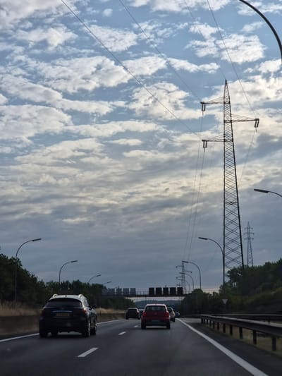 A highway scene under a cloudy sky. The image is oriented vertically, suggesting it might have been taken from a moving vehicle. In the foreground, a dark-colored SUV is on the right lane of the highway, with its license plate visible: "CB 5254". Other cars are visible ahead and behind.
Dominating the left side of the frame are large electricity pylons and power lines against a sky filled with fluffy white clouds. The clouds are wispy and elongated, suggesting a dynamic weather pattern. The overall lighting indicates it is daytime, possibly late afternoon or early morning, given the soft light and the mix of bright and darker clouds.
Streetlights with curved poles line the right side of the highway, and the road itself is wet, hinting at recent rain or damp conditions. Dense green trees and vegetation line the side of the highway. In the distance, more electricity pylons are visible, stretching into the landscape. There are no people clearly visible in the image. The scene appears to be set on a highway in Mondercange, Luxembourg.