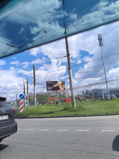 The image shows a roadside view, likely from a moving vehicle, in Малая Валяевка, Russia. In the foreground, the rear of a dark-colored Toyota SUV is visible on the asphalt road, with its license plate reading "11PY 193 RUS". A green grassy verge with a curb separates the road from a sidewalk area.
Further back, several utility poles are spaced along the grassy area. A blue directional sign indicating a left turn is mounted on one of the poles. A billboard with the text "ФОРТ 2 дом" (FORT 2 house) and an image of a woman wearing a wide-brimmed hat is prominently displayed. Text below the title suggests a new collection of apartments and a phone number "500 648".
Beyond the billboard, a series of buildings, including a blue and white structure with large windows, are visible. A tall, white telecommunications tower with multiple antennas stands against a backdrop of a blue sky with scattered white clouds. A plume of dark smoke is rising in the distance, possibly from an industrial source or a fire. A lone person, appearing to be a woman in dark clothing, is walking on the sidewalk.