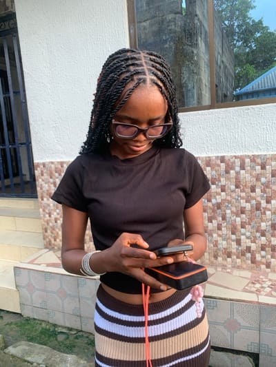 A young woman with braided hair and glasses is standing outside. She is wearing a dark brown shirt and a striped skirt. The woman is holding two phones in her hands. A white wall and window are behind her on the left, and a brick wall is on the right. The ground is tiled.