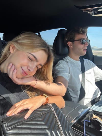 An interior view of a vehicle shows two individuals, a female and a male, in the front seats during daytime. The location is Santarém, Portugal.
The female is seated in the passenger seat on the left, secured by a black seatbelt. She has blonde hair and is depicted with her eyes closed, a soft smile on her face, and her right hand supporting her head. Her left hand rests on a black quilted handbag with a silver chain strap. She wears a dark, ribbed top, a thin red string bracelet on her right wrist, and a gold-toned rectangular watch with a brown leather strap on her left wrist.
The male, in the driver's seat on the right, is also secured by a black seatbelt. He has brown, curly hair and wears dark sunglasses with brown frames. His mouth is slightly open, and he appears to be looking forward, operating the vehicle. He wears a light-colored short-sleeved t-shirt with dark text, which includes phrases like "THE HIDDEN SYMPHONY" and "EXPLORING THE ETHNOBOTANICAL PRINCIPLES IN ART, AND DESIGN."
The car's interior features black upholstery and a dark headliner. A center console is partially visible, showing a blue charging cable connected to an unseen device. Through the windows, a clear blue sky with some distant white clouds and what appears to be a multi-lane road or highway are visible, indicating the vehicle is in motion. Sunlight illuminates the interior.