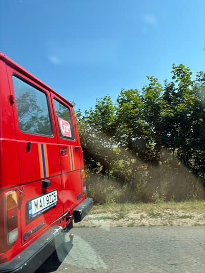 A red van, possibly an emergency vehicle, is parked on the side of a paved road. The van has the number "112" in a red square on its rear window, indicating an emergency services number. The license plate reads "MAI 9225". The van has stripes of blue and orange on its side. Behind the van, there is a line of green trees and vegetation. The sky is clear and blue. The image was taken in the city of Marcăuți, Moldova.