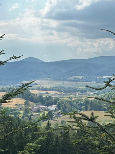This is an eye-level, landscape photograph taken from a high vantage point overlooking a rural area in or near Chełmsko Śląskie, Poland. The foreground is dominated by the dark green branches and needles of coniferous trees, particularly on the left and right sides of the frame, framing the scene. In the midground, a village or small town is visible, with several buildings, including larger structures with light-colored facades and smaller, darker buildings. Fields of varying colors, including golden-brown and green, are interspersed with the trees and buildings. In the background, rolling hills covered in dense green forests recede into the distance, culminating in a hazy, blue mountain peak under a sky filled with dramatic, white and gray clouds. There are no people or animals clearly visible in the scene. The overall impression is of a serene, bucolic landscape bathed in natural light.
