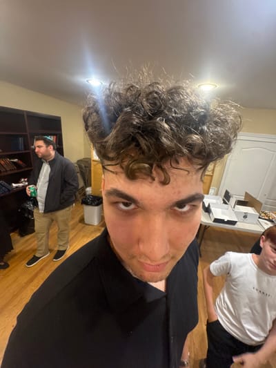 An indoor scene in Bergenfield, United States, depicts three males within a casual gathering space. In the immediate foreground, a young male with voluminous dark curly hair and a black collared shirt looks directly into the camera with an intense expression, his eyebrows slightly furrowed. To the mid-ground left, an adult male wearing a dark yarmulke, a dark jacket over a light collared shirt, and khaki pants stands holding a green soda can in his left hand, looking towards the right side of the frame. Behind him is a dark wooden bookshelf stocked with numerous books, many appearing to be religious texts. To the far background right, a young male with reddish-brown hair and a white t-shirt featuring a dark logo stands with his hands in his pockets, partially visible. The room features light wooden flooring, light beige walls, and a white ceiling equipped with recessed lighting fixtures. A white rectangular table in the background holds multiple open white cardboard boxes, possibly for food, and various baked goods or snacks. A white trash can with a black liner is positioned near the table, and a white door is visible further in the background on the right.