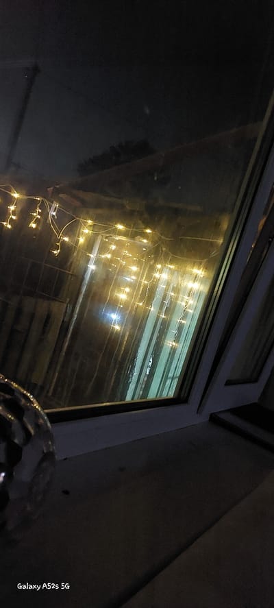 This image, taken at night, shows a window with string lights hanging outside. The lights are yellow and illuminate some sort of structure behind the window, possibly a shed or outbuilding. There is a dark, reflective surface in the foreground, and the window itself appears slightly dirty or wet. The bottom right corner of the image shows the text "Galaxy A52s 5G". The scene is likely a residential or commercial exterior, illuminated by artificial light sources. There are no people visible in the image.