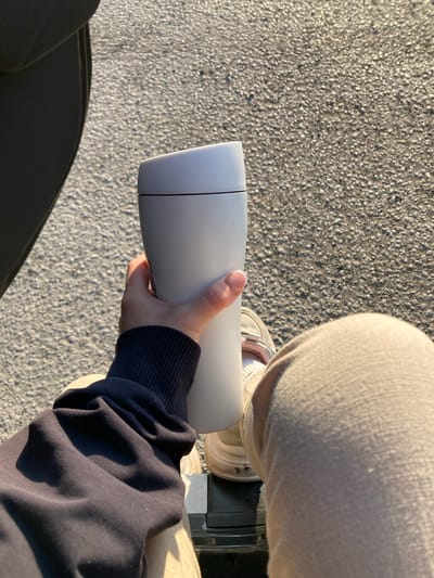 A person is seated, holding a white thermos-like container in their right hand. The person is wearing light-colored pants, white sneakers, and a navy blue sweater. The background consists of an asphalt surface and parts of what appear to be a wheelchair. Verkh-Sosnova, Russia, is not evident from the provided image.