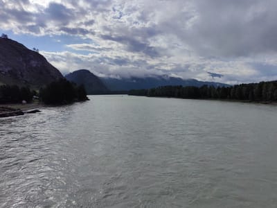 This is a wide shot of a wide, grey river flowing through a valley in Anos, Russia. The river takes up most of the frame, with choppy water creating ripples and reflections. On the left side of the river, a steep, tree-covered mountain rises sharply, with a small patch of bare ground at its base. On the right side of the river, a dense line of dark green trees forms a natural border, with more mountains visible in the distance, partially obscured by clouds. The sky above is a dramatic mix of grey and white clouds, with patches of blue sky peeking through. There are no people or specific structures visible in the scene.