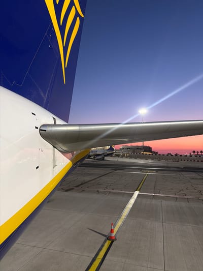 The image shows part of a Ryanair aircraft at Malta International Airport in Luqa, Malta. The tail of the aircraft is visible with the Ryanair logo in yellow on a blue background. The white wing with a yellow stripe is in the foreground, extending towards a grey concrete tarmac. A street lamp is visible above the plane, and a cone and a yellow line are seen in the background. There is a sunset with orange and pink hues in the background. Buildings and palm trees can be seen in the distance.