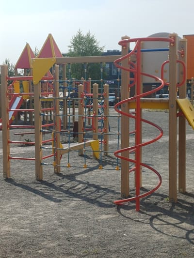 This is a low-angle, vertically oriented photograph of a modern, multi-level outdoor playground structure. The playground features a variety of climbing elements, including a rope net, what appears to be a spiral slide or climbing structure made of red metal, and wooden beams serving as steps or platforms. Colorful yellow and pink triangular elements are integrated into the structure, possibly as roofs or decorative features. The main materials are wood, red metal, and blue rope.
The playground is situated on a gravel surface, with shadows of the structure cast upon it, suggesting it is daytime. The sky is visible at the top of the frame, indicating an outdoor setting under natural light. In the background, there are hints of trees and buildings, including what looks like a modern commercial building with windows. One visible sign contains partial text, appearing to read "NOZ".
No people are present in the image, and the scene appears to be a quiet, unoccupied playground. The time of day seems to be late morning or afternoon, given the length and angle of the shadows. The weather appears to be clear and sunny. The overall impression is one of a well-maintained, contemporary recreational facility.