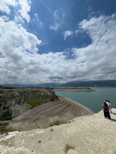 Two individuals are positioned on the edge of a steep cliff in Chirkey, Russia. The cliff overlooks a body of water and a landscape featuring layered geological formations. The sky is partially covered with white clouds.