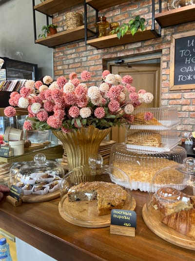 The image depicts a cafe counter in Adzhiryuara, Abkhazia, displaying a variety of baked goods and floral arrangements. In the foreground, a wooden counter holds several items under glass domes and in plastic containers. These include individual pastries, a whole cake, and sliced cakes, likely Napoleon cake given the visible layers. A sign with Cyrillic text and the price "160" is placed in front of a slice of cake. To the left, a hand reaches towards a display of small chocolate-covered pastries, also with a Cyrillic label and price "250". Behind these, a large vase overflows with pink and white faux flowers.
The background features a brick wall with shelves holding decorative items such as woven baskets and glass vases. A blackboard with chalk writing is also visible on the wall. A wooden door is present in the center of the background. The overall environment suggests a retail setting focused on food service, with a rustic yet organized aesthetic.