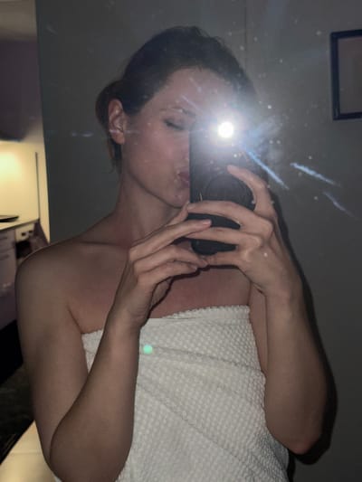 A woman is taking a selfie in a mirror. She is holding a phone in front of her face, partially obscuring it. She is wrapped in a white towel. A framed picture is visible in the upper left corner of the mirror. The location is not identifiable as Kranj, Slovenia.