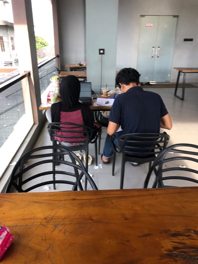 In Geulumpangsulu Timu, Indonesia, two individuals are seated at tables in an outdoor cafe or communal workspace. On the left, a person wearing a black hijab and a maroon top is facing away from the camera, looking at a laptop on a table. Next to the laptop are a drink with a straw and other miscellaneous items. To the right, a person wearing a dark blue polo shirt and blue jeans is also seated, facing away from the camera and looking down at a table, possibly working or studying. They are seated at a separate table from the first individual. The environment appears to be an open-air space with tables and chairs, with a building visible in the background and a balcony railing.