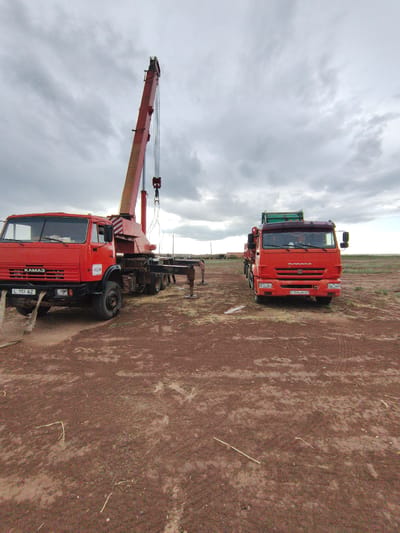 In the settlement of Mergen rural district, Kazakhstan, two red Kamaz trucks are parked on a dirt ground under a cloudy sky. The truck on the left has a crane attached, its boom extended upwards. The truck on the right appears to be a flatbed truck with a green structure on its rear. The ground is reddish-brown and uneven, with sparse dry grass and some scattered debris. In the background, the landscape is flat and appears to be a rural or semi-arid area, with some distant structures visible on the horizon. There are no people clearly visible in the immediate vicinity of the trucks. The overall scene suggests a work or construction site in an open, undeveloped area.