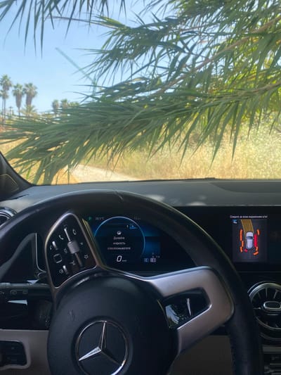 The image shows the interior of a car, specifically the dashboard and steering wheel, as viewed from the driver's seat. Outside the car, through the windshield, a sunny, arid landscape with palm trees is visible, suggesting a warm climate. The scene appears to be in Bel Air, Spain.
The dashboard displays several indicators. On the right side, a parking assist system is active, showing a graphical representation of the car and its surroundings with proximity warnings. On the left side of the dashboard, a digital display shows the current temperature as 28.5°C and a message in Russian that translates to "Top up windshield washer fluid". Below this, the speed is indicated as 0 km/h, suggesting the car is stationary.
The steering wheel is a prominent object, featuring the Mercedes-Benz logo in the center. Various controls are visible on the steering wheel, including buttons for media playback, phone calls, and cruise control. The overall impression is that of a car parked in a sunny, possibly rural or suburban, location in Spain on a clear day. No people are visible in the image.