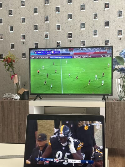 The image depicts an indoor environment with a textured grey wall densely covered by numerous small, square photographs. A large television screen, centrally positioned on a white and wooden media console, displays a FIFA U-17 Women's World Cup soccer match between Mexico and Italy, with a 0-0 score at the 89th minute. On the console's left, a clear glass vase holds a multi-colored floral arrangement with a pink ribbon, alongside white coiled cables and reading glasses. On the right, a second clear vase contains blue artificial flowers. In the foreground, a laptop screen displays an American football game, showing a player in a black and yellow uniform, number 95, from a team with a score of 27 points against an opponent with 10 points in the fourth quarter. No individuals are physically present within the depicted room. The image provides no discernible visual information to confirm a specific location within Santa Catarina, Mexico.