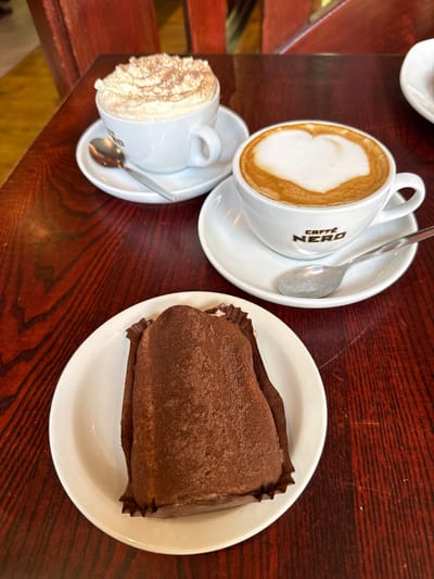 The image shows a cafe setting on a dark wood-grained table. There is a caffe latte in a Caffe Nero branded cup with a spoon on the saucer, and a second cup of beverage covered in whipped cream with a spoon on its saucer. Also on the table, sitting on a white plate, is a long rectangular, brown cake covered in cocoa powder, still in its crinkled paper wrap. This scene is consistent with a caffeinated coffee and pastry cafe location, for example Caffe Nero, or a similar cafe located in the city of Leicester, United Kingdom.