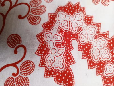 The image displays a close-up view of a patterned fabric, likely cotton or a similar material. The dominant colors are red and white. Red floral designs with white flower-like accents are repeated across the surface. The red sections feature dotted patterns, adding texture and visual interest. The white background provides contrast, enhancing the vibrancy of the red motifs. The overall impression is that of a traditional or folk-inspired textile design.