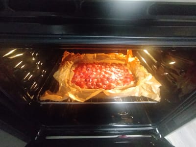 A rectangular pastry, possibly a tart or pie, sits on a baking sheet lined with parchment paper within an open oven. The pastry is filled with what appears to be chopped strawberries or another red fruit, glistening under the oven light. The pastry itself has a light brown, possibly golden-brown crust, which is partially obscured by the crumpled parchment paper. The oven interior is dark, with its light illuminating the pastry and reflecting off the oven racks and walls. The scene suggests baking is in progress. There is no visible text in the image.
