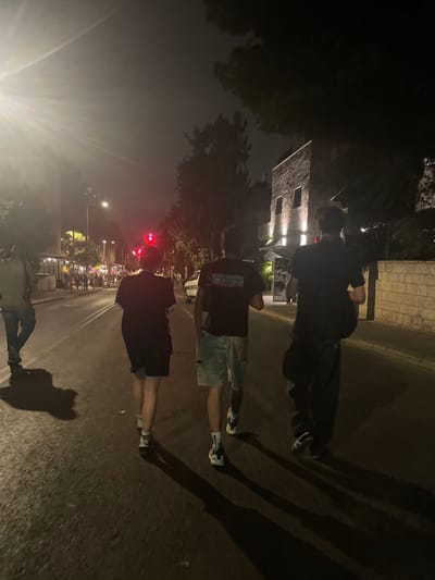 The image captures a nighttime street scene in West Jerusalem, Israel. Four individuals are walking away from the camera on an asphalt road. The closest subject on the far left is partially visible, wearing a light-colored hooded garment and dark pants. Two central subjects walk abreast: one in a dark t-shirt and dark shorts, and the other in a dark t-shirt with "FAMILIES FORUM" visible on the back, light denim shorts, and white sneakers. The fourth subject on the far right wears a dark t-shirt, dark pants, and carries a dark shoulder bag. Long shadows from the individuals are cast on the road due to a bright streetlight flaring from the upper left. In the mid-distance, a red traffic light is visible. To the right, a multi-story building constructed of light-colored stone blocks features facade lighting. Trees line the street, and a white van is partially visible further down the road on the right. In the far distance, multiple lights illuminate a gathering of people and structures, suggesting ongoing activity.