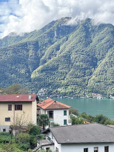 This image presents a panoramic view of the landscape in Nesso, Italy, specifically showcasing a section of Lake Como and its mountainous surroundings.
In the foreground, an arrangement of residential buildings descends a slope. The most prominent structures include a two-story beige building with a terracotta tiled roof, brown window frames, and partially visible dark brown shutters. To its right, another white building with a terracotta tiled roof features green shutters on its windows and a balcony opening. Further down and partially obscured, a third, smaller white building with a dark grey tiled roof is visible, featuring an external concrete staircase with railings leading to an upper landing. These buildings are interspersed with dense green vegetation, including trees and shrubs. No individuals are present or visible in this section.
The midground is dominated by a wide expanse of teal-colored water, identified as Lake Como, exhibiting subtle ripples across its surface. Along the distant shoreline, numerous light-colored residential and commercial structures are clustered at the base of the mountains.
The background is characterized by a series of steep, imposing mountains heavily covered in dense green foliage, indicating extensive tree growth. The upper slopes and peaks of these mountains are partially obscured by white and light grey clouds. The sky above is partly cloudy, with patches of blue sky visible between the white and grey cloud formations. The scene is illuminated by natural daylight.