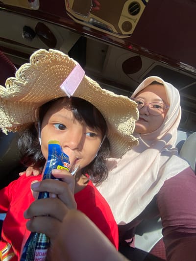 Within Sumber Suko, Indonesia, an interior scene captures two females, likely inside a vehicle such as a bus or large van, illuminated by strong sunlight from the left, creating harsh shadows. In the foreground, a young child, approximately 5-10 years old, with dark hair and medium skin tone, is wearing a light-colored woven straw hat adorned with a pink ribbon and a red t-shirt. She holds a blue and white flexible package labeled "Singa," "KEJU," "2X LEBIH," and "UDANG GORENG," containing a green substance, to her mouth, actively consuming its contents. Her gaze is directed slightly to her right. Behind and to the right, an adult female, likely in her 20s or 30s, with medium skin tone, wears a light pink or beige hijab and purple-framed eyeglasses. She is dressed in a maroon long-sleeved top. Her right hand rests gently on the child's left shoulder. Both individuals are looking towards the camera. The background reveals overhead panels, air vents, and lighting fixtures characteristic of a vehicle interior, with dark upholstery visible on the seats.