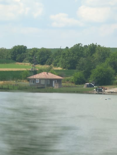 This is a vertically oriented, landscape-oriented photograph taken from a slight elevation. The scene depicts a rural setting in Maslarevo, Bulgaria. In the foreground, a large body of water, likely a lake or river, occupies the right half of the frame, its surface rippled and reflecting the surrounding environment with a muted green hue.
To the left of the water, a narrow strip of land contains a low-slung building with a reddish-brown tiled roof and dark grey walls. The building appears to have several windows and a white door. A prominent logo featuring the letters "BK" in white within a dark rectangular frame is visible on one of the walls. Adjacent to the building, a dark, abstract structure, possibly a piece of equipment or an antenna, stands tall.
Beyond the building, a vibrant green field stretches out, dotted with trees. Further in the background, a dense line of trees forms a dark green band against a bright blue sky scattered with fluffy white clouds. The overall impression is of a peaceful, sunny day in the countryside. There are no people visible in the image. The orientation of the image suggests it may have been taken from a moving vehicle or a high vantage point.