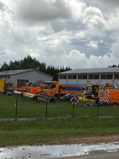 In Odukalns, Latvia, an outdoor scene features a collection of specialized vehicles, possibly for road maintenance or snow removal, parked on a grassy area in front of a long, low-rise industrial building with a grey roof and a row of windows. The vehicles are predominantly orange and yellow, with some featuring large plows or salt spreaders. Among them are at least two large trucks, one with a visible Mercedes-Benz logo, and a yellow tractor or loader. Several traffic signs, including those with orange and white diagonal stripes and blue circular signs with white symbols, are visible near some of the vehicles, suggesting active use or preparation for operation. The ground in the foreground is wet and appears to have large puddles, indicating recent rain. The sky is overcast with dramatic, gray clouds. The environment suggests a depot or yard for heavy machinery. There are no people visible in the image.