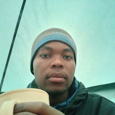 An adult male with a dark complexion is depicted in a close-up, likely a selfie, from inside a tent at Gilman's Point, Tanzania. He is wearing a grey and blue striped beanie and a dark jacket with a blue inner collar. His gaze is directed towards the camera, and his expression is neutral. He holds a light-colored mug, possibly cream or light yellow, in his right hand, which is partially visible in the lower left foreground. The background is composed of a teal-colored fabric, indicative of a tent interior, with a dark vertical line, possibly a tent pole or seam, visible on the left.
